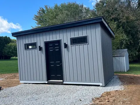 Gray outbuilding with vertical siding, a black door and a gravel base